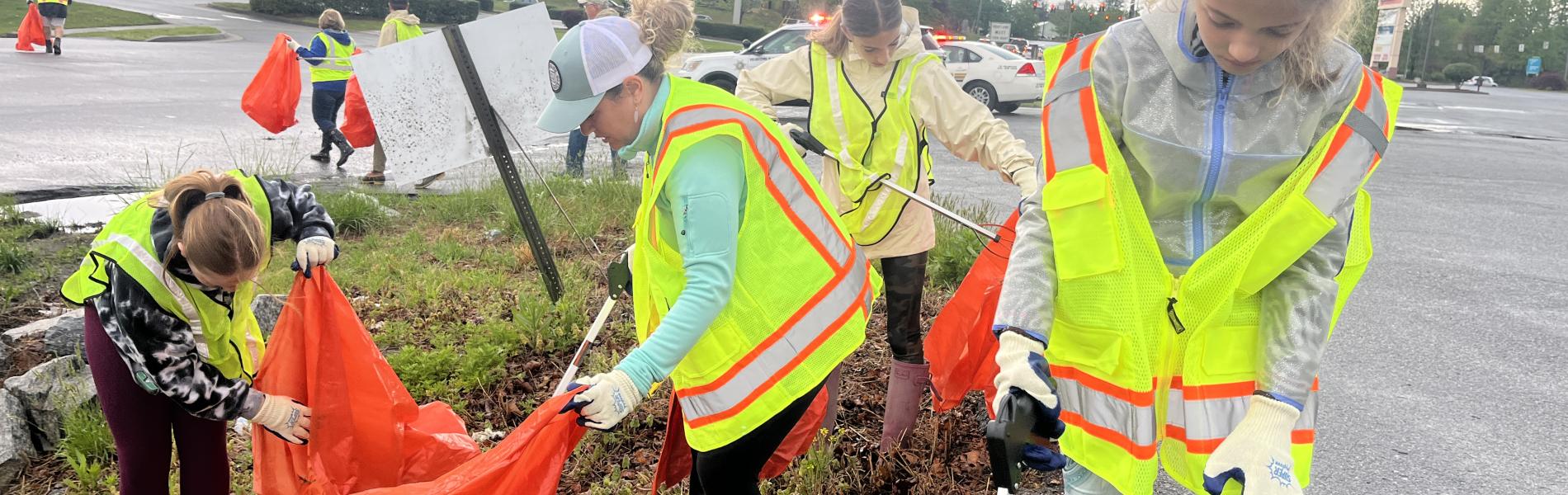 A group of people wearing yellow vests, picking up litter and disposing in orange bags.