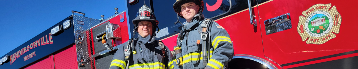 Two men in front of fire engine wearing firefighter gear.