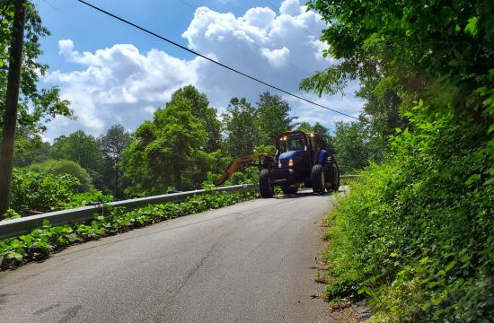 Sidearm mower driving down the street, with green vegetation growing on both sides of the road.