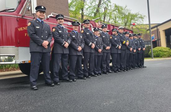 A large group of Firefighters in their uniforms stand in front of a red fire engine.