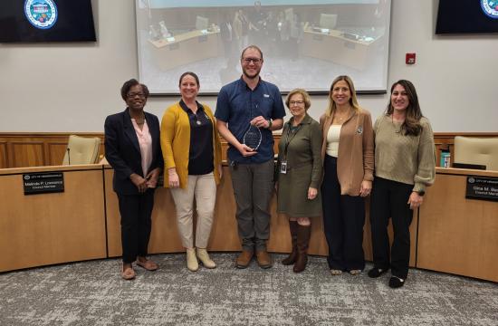 Six people stand together smiling, with the person in the middle holding an award.