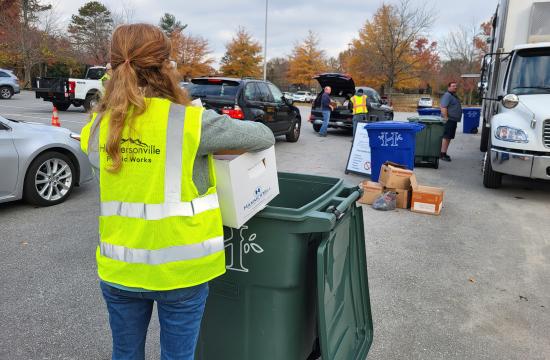 Employee in yellow safety vest dumps documents to be shredded in a green trash container at public shred event.