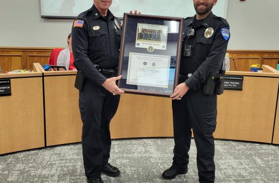 Two police officers stand in a conference room, holding a framed certificate.