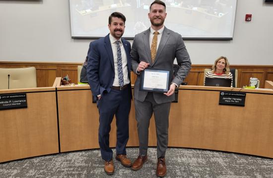 Two people stand in a meeting room, one holds a certificate and both are smiling.