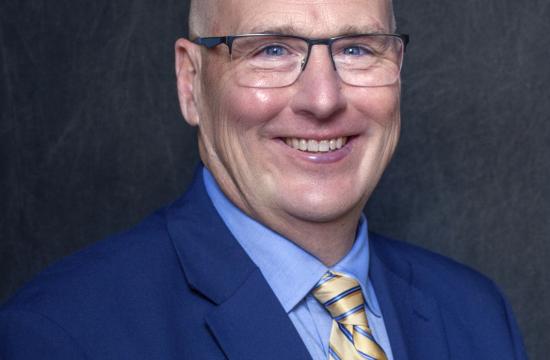 A headshot of a man in a dark blue suit, wearing glasses and a yellow tie smiling