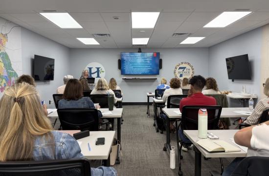 Conference room with people sitting at tables, actively listening to a presenter up front.