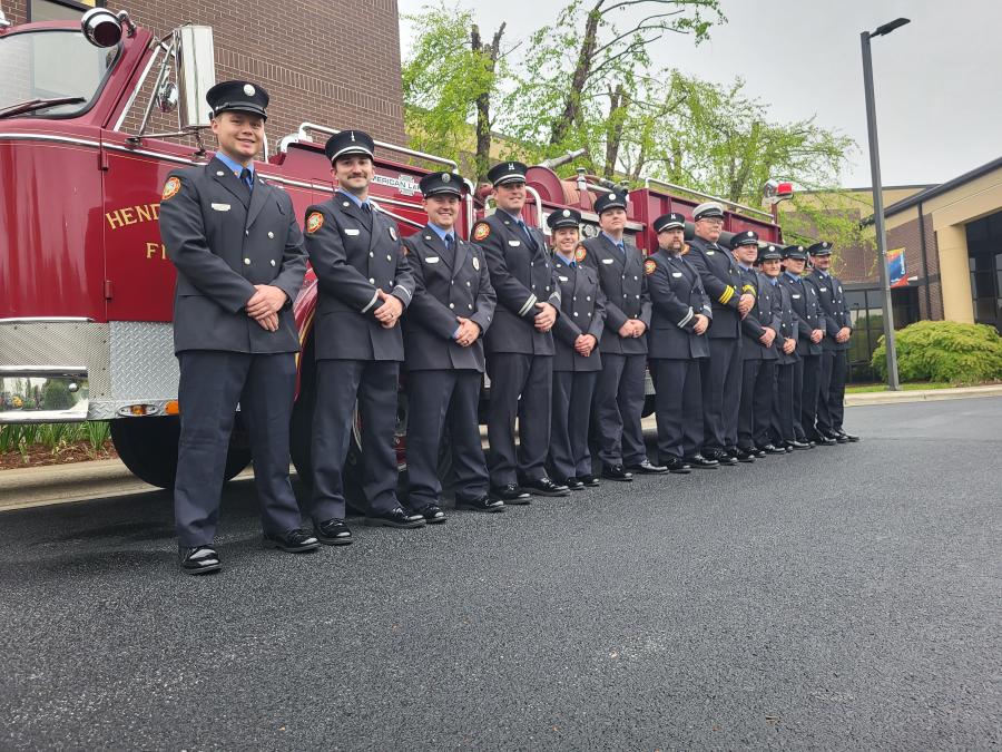 A large group of Firefighters in their uniforms stand in front of a red fire engine.