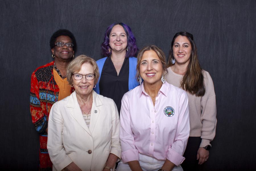 Five people stand together smiling, with a dark gray backdrop behind them.