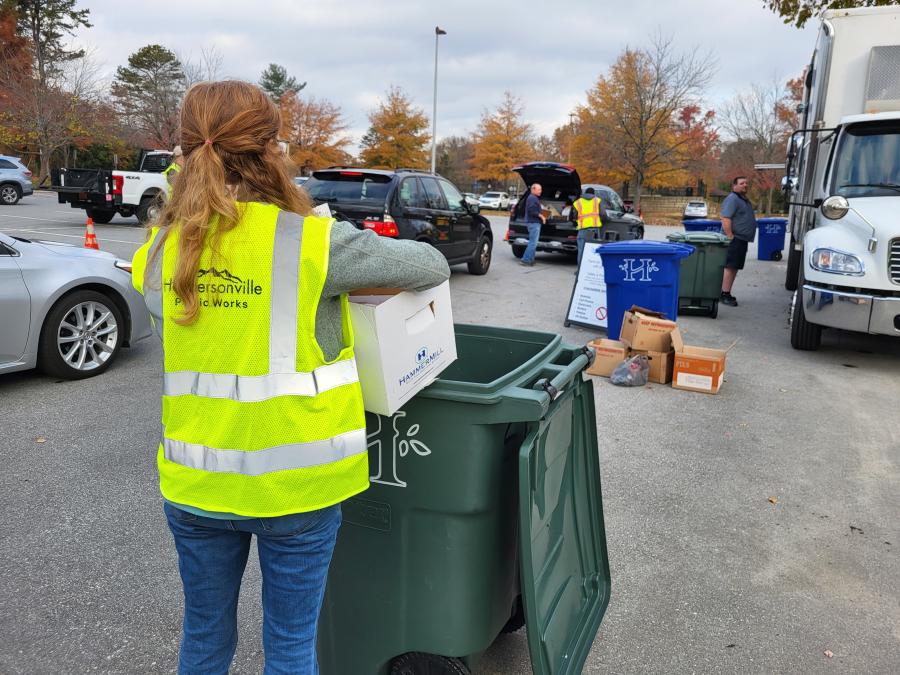 Employee in yellow safety vest dumps documents to be shredded in a green trash container at public shred event.