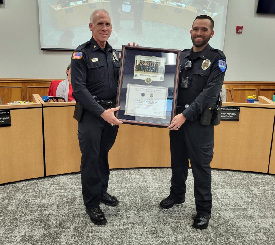 Two police officers stand in a conference room, holding a framed certificate.