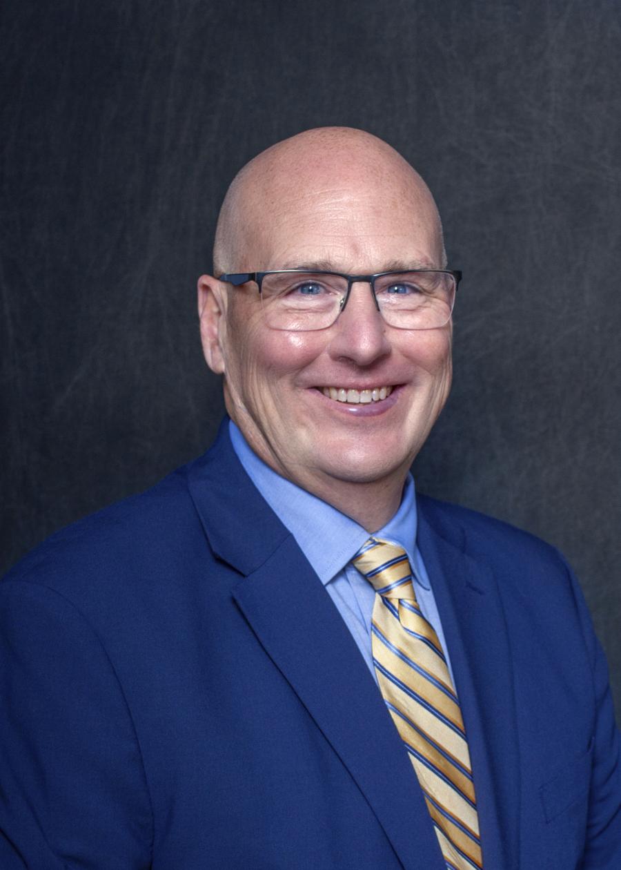 A headshot of a man in a dark blue suit, wearing glasses and a yellow tie smiling