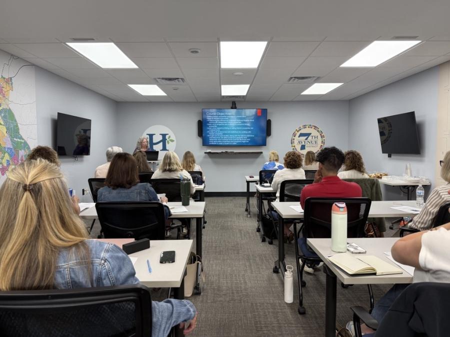 Conference room with people sitting at tables, actively listening to a presenter up front.