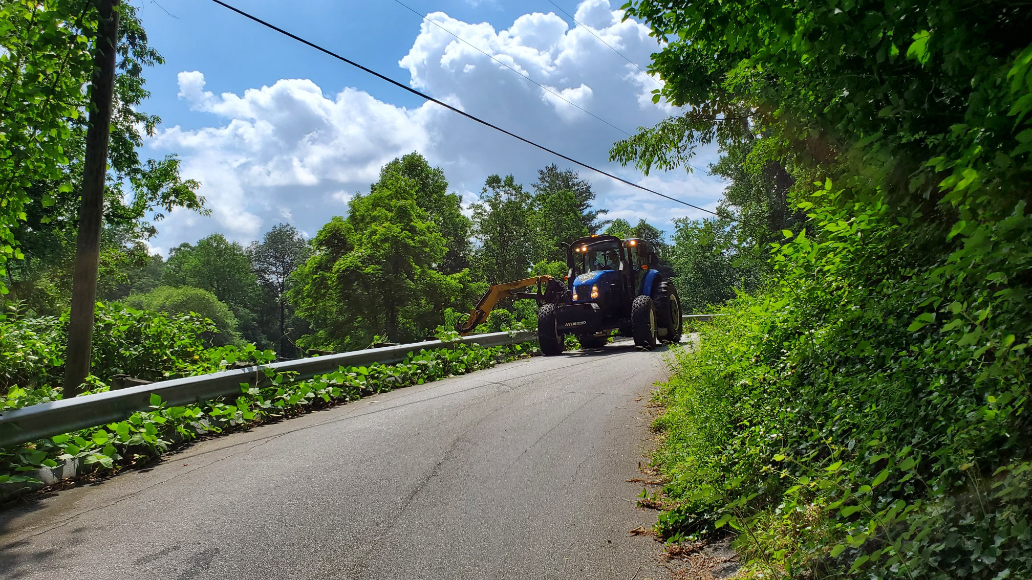 A large piece of Public Works equipment with an arm cuts excessive greenery around a guardrail on a street.