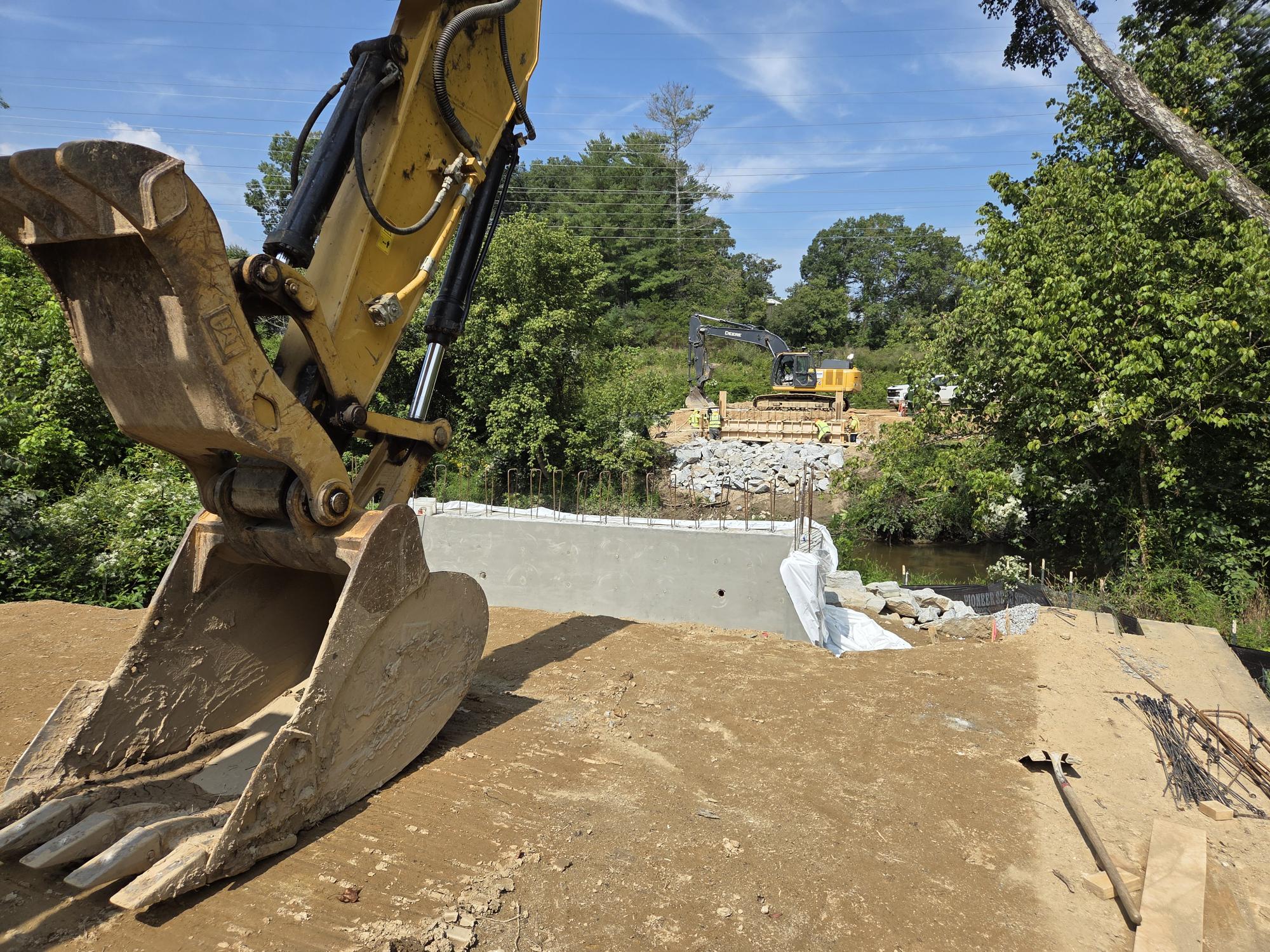 Photo looking across Mud Creek. Foreground is a backhoe bucket resting on the ground and a concrete bridge abutment.